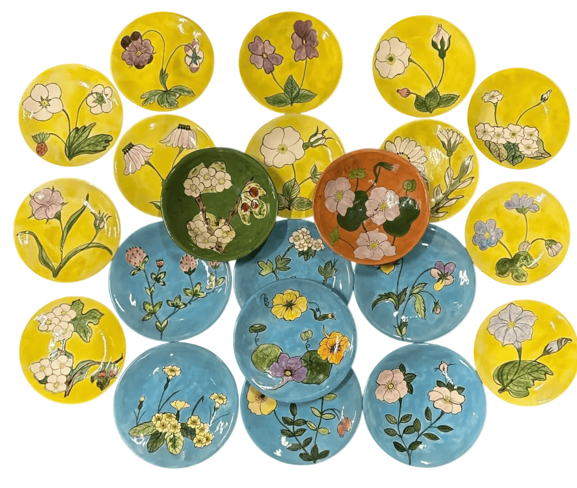 A collection of round ceramic plates with hand-painted floral designs, featuring bright yellow, light blue, green, and terracotta backgrounds with white, pink, and purple flowers.