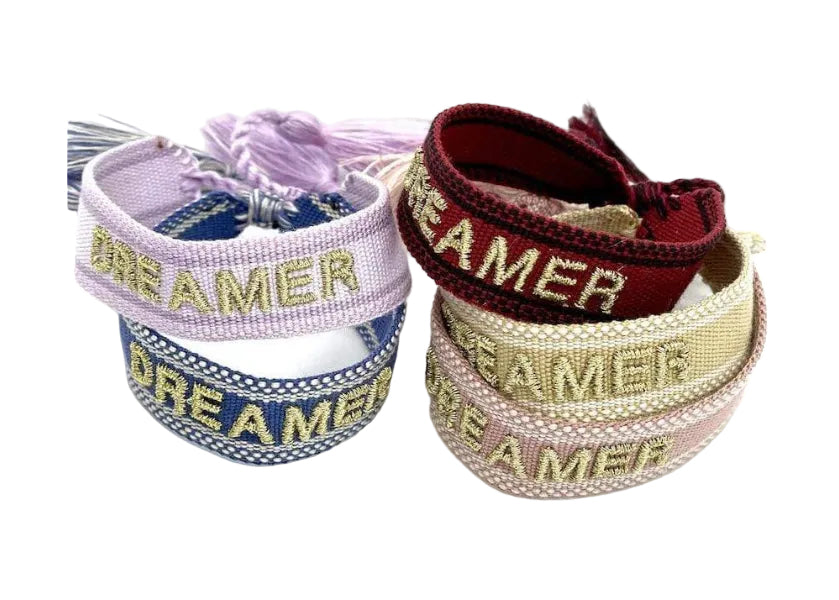 A collection of woven cotton friendship bracelets with adjustable straps. Each bracelet features the word 'DREAMER' embroidered on it, available in different colors such as pink, blue, red, beige, and green.